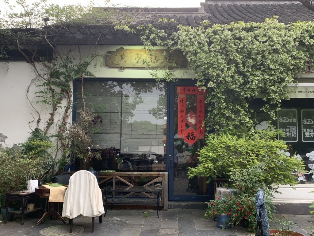Un petit salon de thé caché dans le coin européen du village de Wangshan à Suzhou. Devanture avec porte bleu foncé et rouge, entouré de verdures et de plantes. Une table se trouve à l'extérieur. 