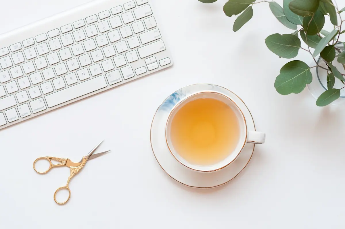 Vue de dessus d'un espace de travail minimaliste avec un clavier blanc, une tasse de thé chaud sur une soucoupe, et un petit vase de feuilles d'eucalyptus.
