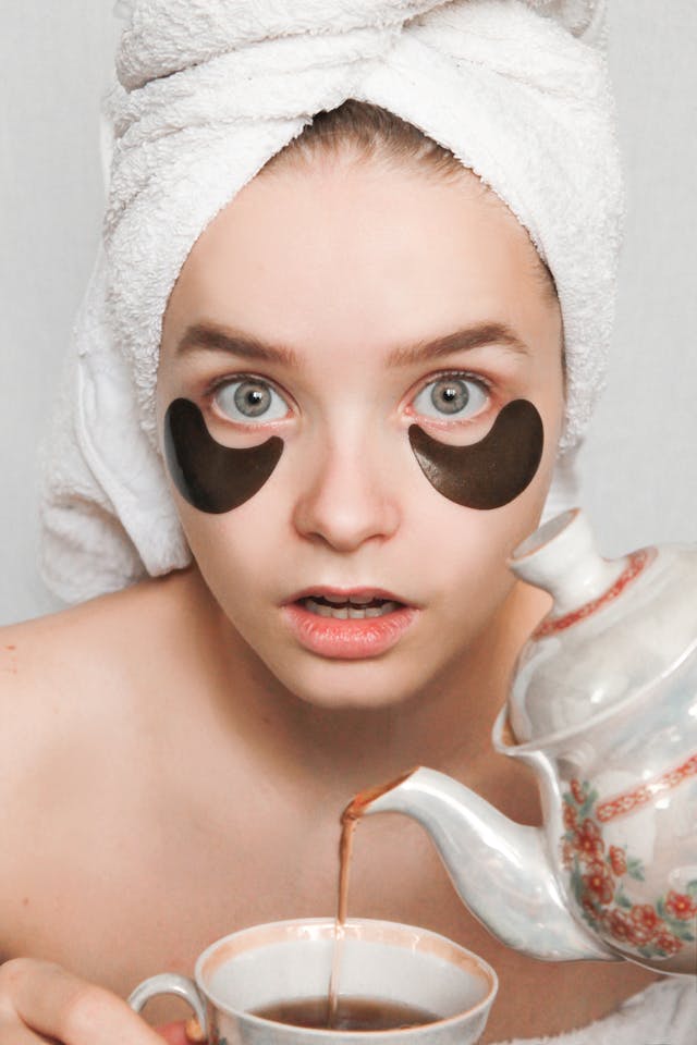 Une jeune femme portant des patchs pour les yeux et une serviette sur la tête verse du thé, illustrant une routine beauté naturelle et les bienfaits du thé sur la peau pour un teint éclatant et lumineux.