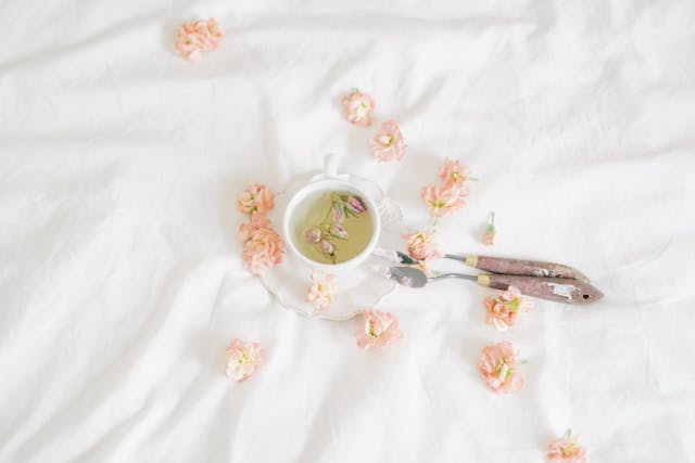 Une tasse d'infusion aux fleurs et boutons de rose disposée sur un drap blanc, illustrant une routine beauté naturelle et les bienfaits du thé sur la peau, comme l'utilisation du thé vert contre l'acné pour un teint éclatant et lumineux.