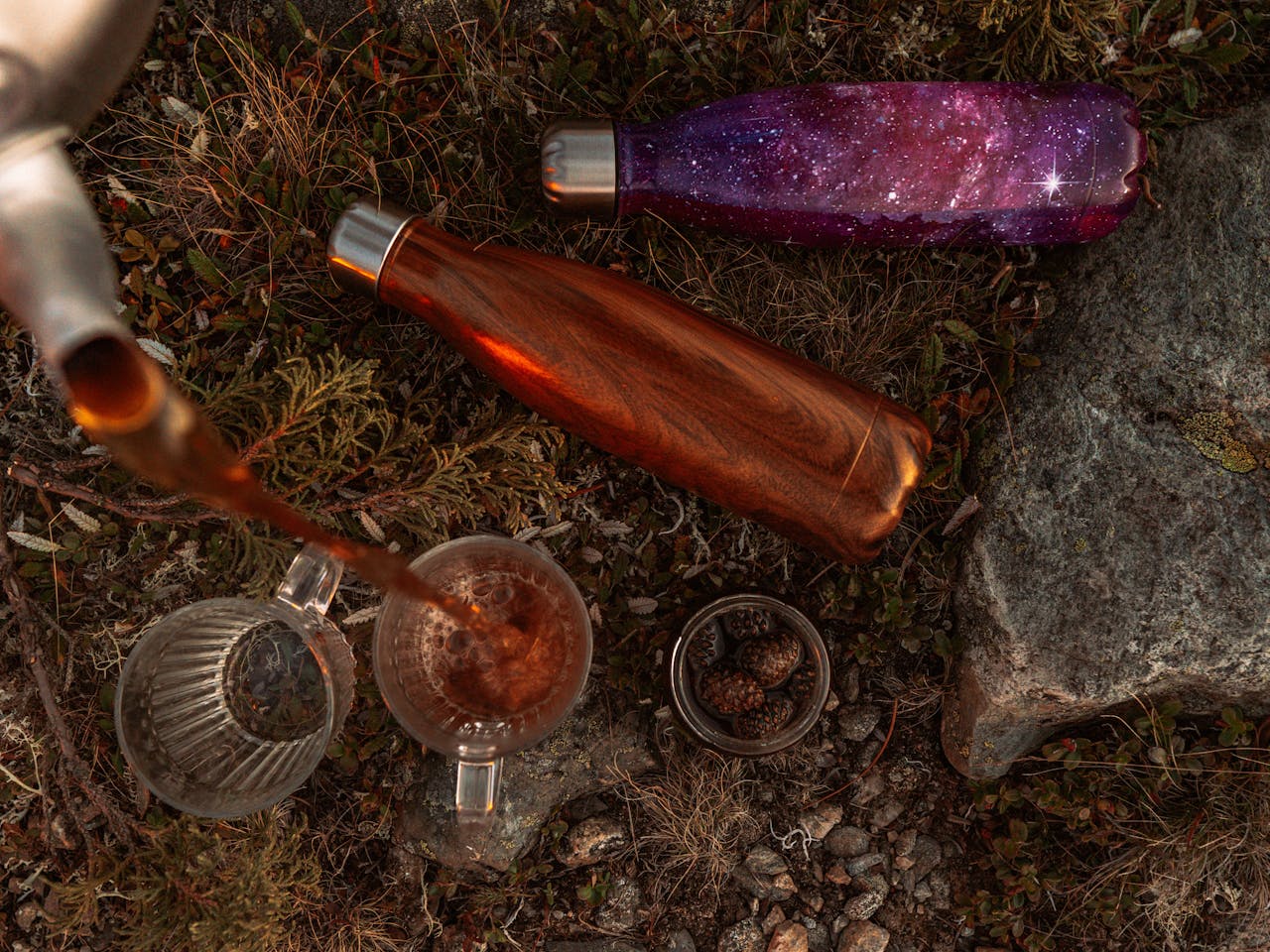 Deux gourdes thermos voyage (rouge cuivré et violet métallique) posées sur l'herbe, à côté d'une théière en verre et de petites tasses. Une invitation à boire du thé en voyage en pleine nature, entre amis. L'esprit thé nomade dans toute sa douceur.