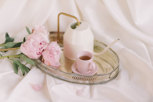 Un plateau doré élégant avec une tasse de thé rose et un arrosoir moderne, posé sur un drap de satin blanc soyeux avec des pivoines fraîches, évoquant une routine de soin et les bienfaits du thé sur la peau pour un teint éclatant et lumineux.