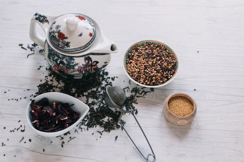 Une théière chinoise en porcelaine entourée de feuilles de thé en vrac, de fleurs d'hibiscus séchées, d'un infuseur à pince et d'un bol de sucre roux sur une table en bois clair.