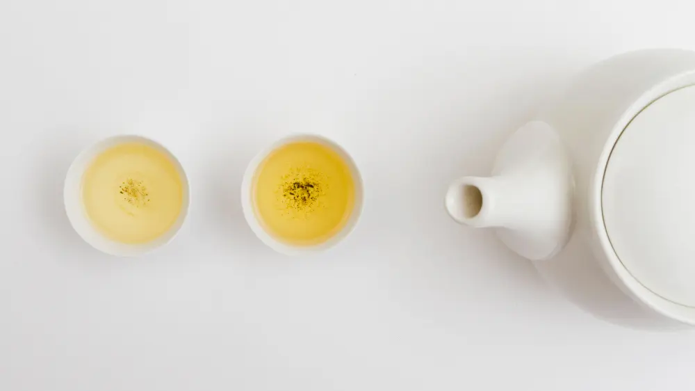 Théière de thé blanche sur un fond blanc, à côté 2 tasses blanches, une de thé blanc et une de thé vert au jasmin, deux thés à boire quand il fait chaud. 