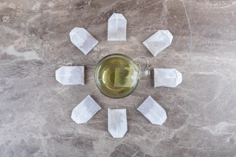 Une tasse de thé infusé entourée de plusieurs sachets disposés en cercle sur une table en marbre, illustrant le dilemme entre thé en vrac et thé en sachets.