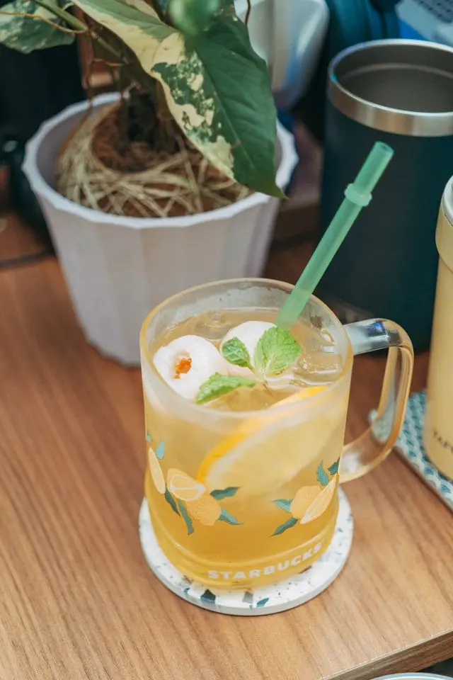 Une tasse en verre Starbucks contenant un thé blanc aux fruits infusé au litchi et au citron avec une paille verte, posée sur un bureau.