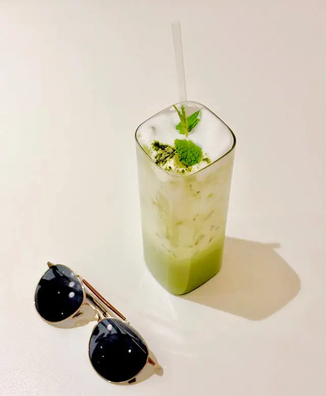 Le meilleur thé quand il fait chaud est le matcha glacé latte dans un verre avec des feuilles de menthe dessus. A côté sont posées des lunettes de soleil. 