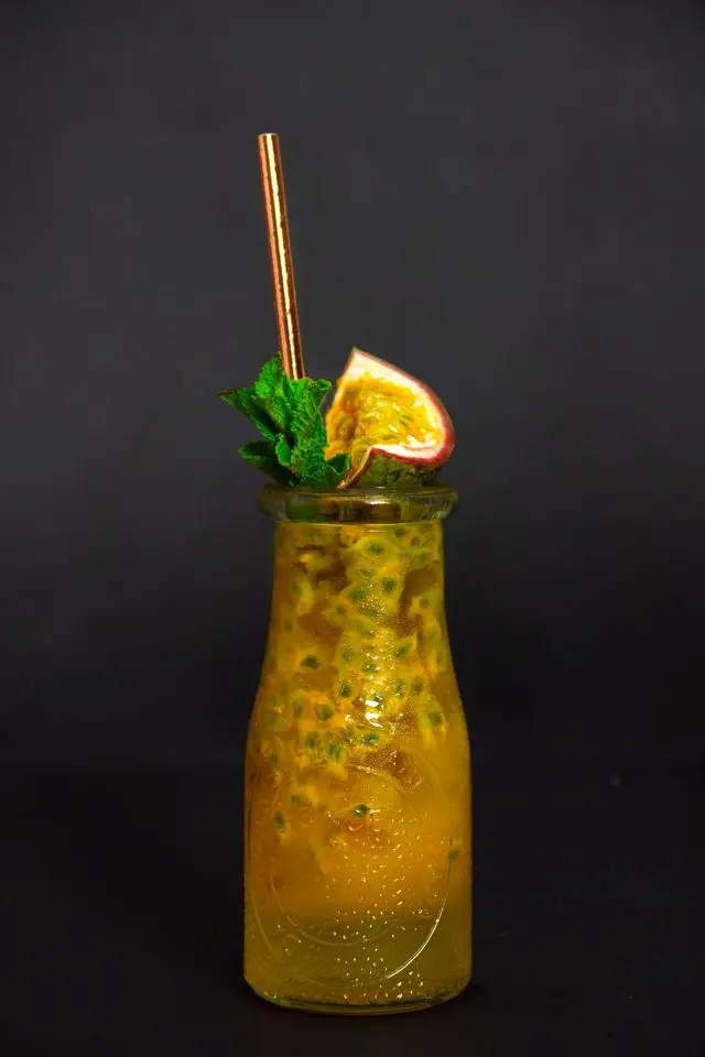 Une bouteille en verre remplie d'un délicieux thé noir aux fruits de la passion avec des graines visibles et une tranche de fruit frais sur fond noir.