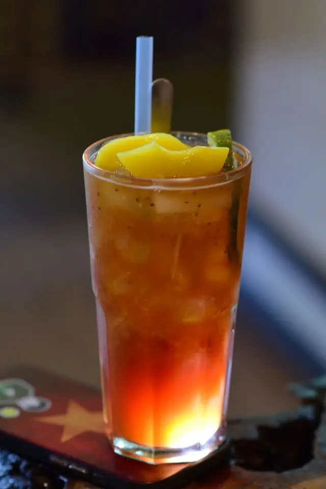 Un grand verre de thé vert glacé dégradé avec des morceaux de fruits tropicaux type mangue sur le dessus, parfait pour illustrer les thés fruités de la saison estivale.