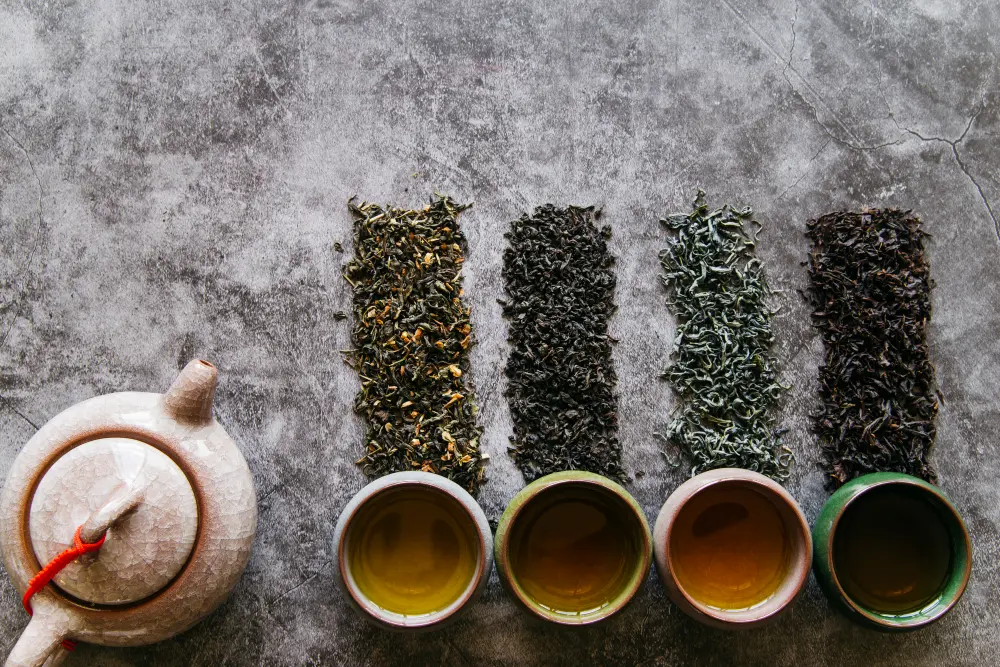 Une vue de dessus de quatre variétés de feuilles de thé sec disposées en colonnes sur un fond en pierre grise texturée. En dessous de chaque sélection, une tasse assortie dévoile une couleur de liqueur différente. Une théière artisanale en céramique craquelée est posée sur la gauche. Comment réussir le thé vert chinois.