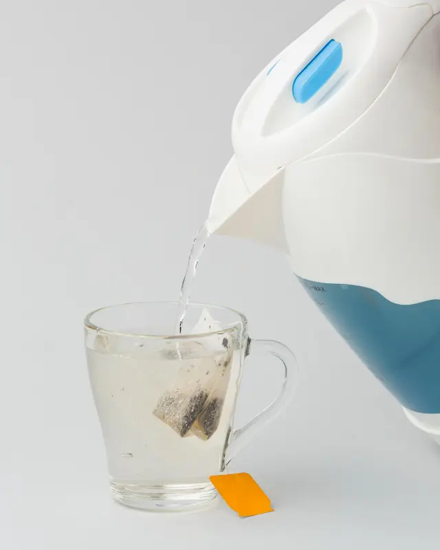 Une bouilloire électrique blanche et bleue verse de l'eau chaude dans une tasse en verre transparent contenant un sachet de thé. Une étiquette orange vif dépasse de la tasse, sur un fond gris clair uni.