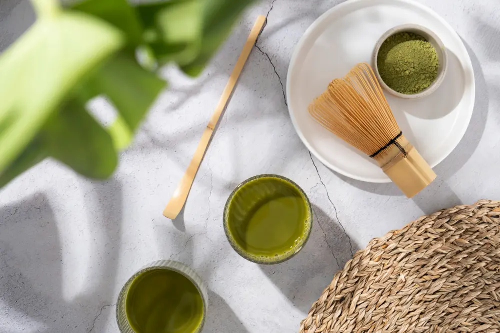 Poudre de thé matcha vert, fouet en bambou et verres d'infusion sur un fond épuré, idéal pour savoir quel thé boire le matin pour un rituel focus et éclat de la peau.