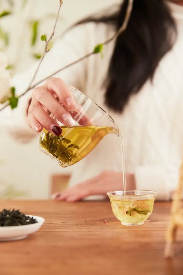 Une femme verse délicatement une liqueur dorée depuis un pichet en verre transparent, contenant des feuilles infusées, vers une petite tasse assortie posée sur une table en bois. Une petite coupelle blanche remplie de feuilles sèches est visible au premier plan. Comment réussir le thé vert chinois et préparer au mieux votre thé vert chinois Biluochun.