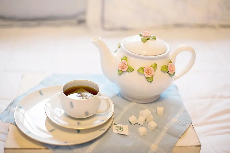 Une tasse de thé fumante avec son sachet et une théière en porcelaine ornée de roses sont posées sur un lit drapé d'un linge bleu, suggérant un moment de détente au réveil pour savoir quel thé boire le matin.