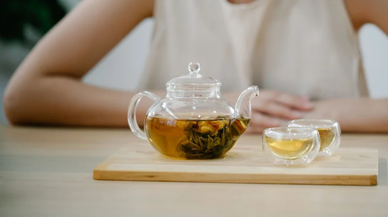 Une théière en verre transparent posée sur une planche en bois, révélant une fleur de thé vert sculptée qui s'épanouit dans une liqueur dorée. À côté, deux petites tasses en verre à double paroi sont remplies d'infusion, avec une personne assise en arrière-plan flou. Comment réussir le thé vert chinois.