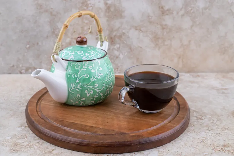 Accessoires pour préparer le thé disposés sur un plateau en bois rond, comprenant une théière en céramique verte à motifs floraux avec une anse en bambou, ainsi qu'une tasse en verre transparent remplie d'un thé sombre, sur un fond en marbre clair.