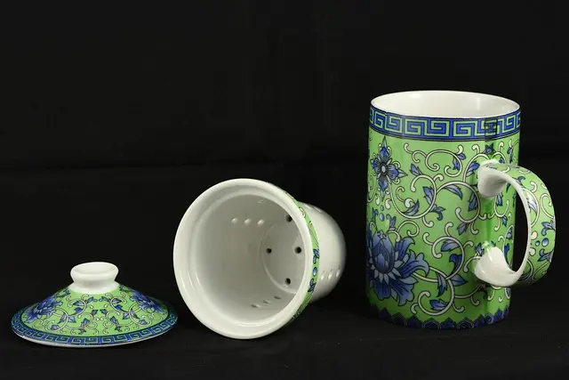 Ensemble de mug à thé en porcelaine chinoise verte et bleue à motifs floraux, comprenant accessoires pour préparer le thé : une grande tasse avec anse, un filtre/infuseur amovible perforé et un couvercle assorti, posés sur un fond noir uni.