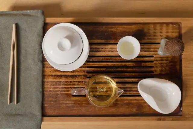 Vue de dessus de matériel pour préparer le thé chinois disposé sur un plateau de service en bois rainuré. L'ensemble comprend un gaiwan en porcelaine blanche avec couvercle, un petit verre de service avec poignée contenant du thé infusé de couleur jaune ambré, une petite tasse à dégustation blanche contenant du thé infusé clair, un repose-théière en céramique blanche et un petit outil à thé en bois posé sur un chiffon gris sur la gauche. L'arrière-plan est un plancher en bois plus clair.