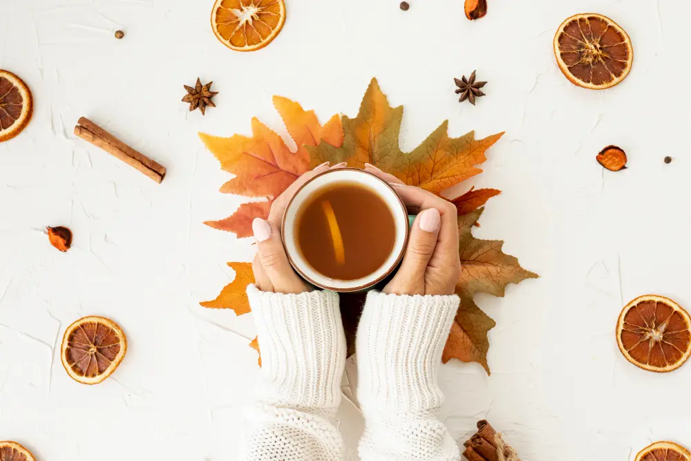 Mains en pull blanc tenant une tasse de thé réchauffant au citron, posée sur des feuilles d'automne avec tranches d'oranges séchées, anis étoilé et cannelle.