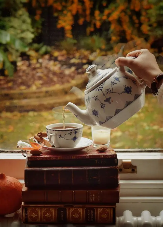 Main versant un thé d'une théière en porcelaine fleurie dans une tasse posée sur une pile de livres anciens près d'une fenêtre en automne.