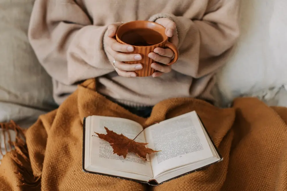 Personne en pull beige tenant une tasse de thé chaud terracotta au-dessus d'un livre ouvert et d'un plaid d'automne en laine.