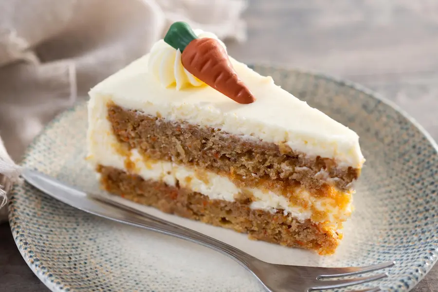 Une part de carrot cake moelleux avec glaçage au fromage à la crème et une petite carotte en sucre décorative, présentée sur une assiette pour un accord thé dessert gourmand cet automne.