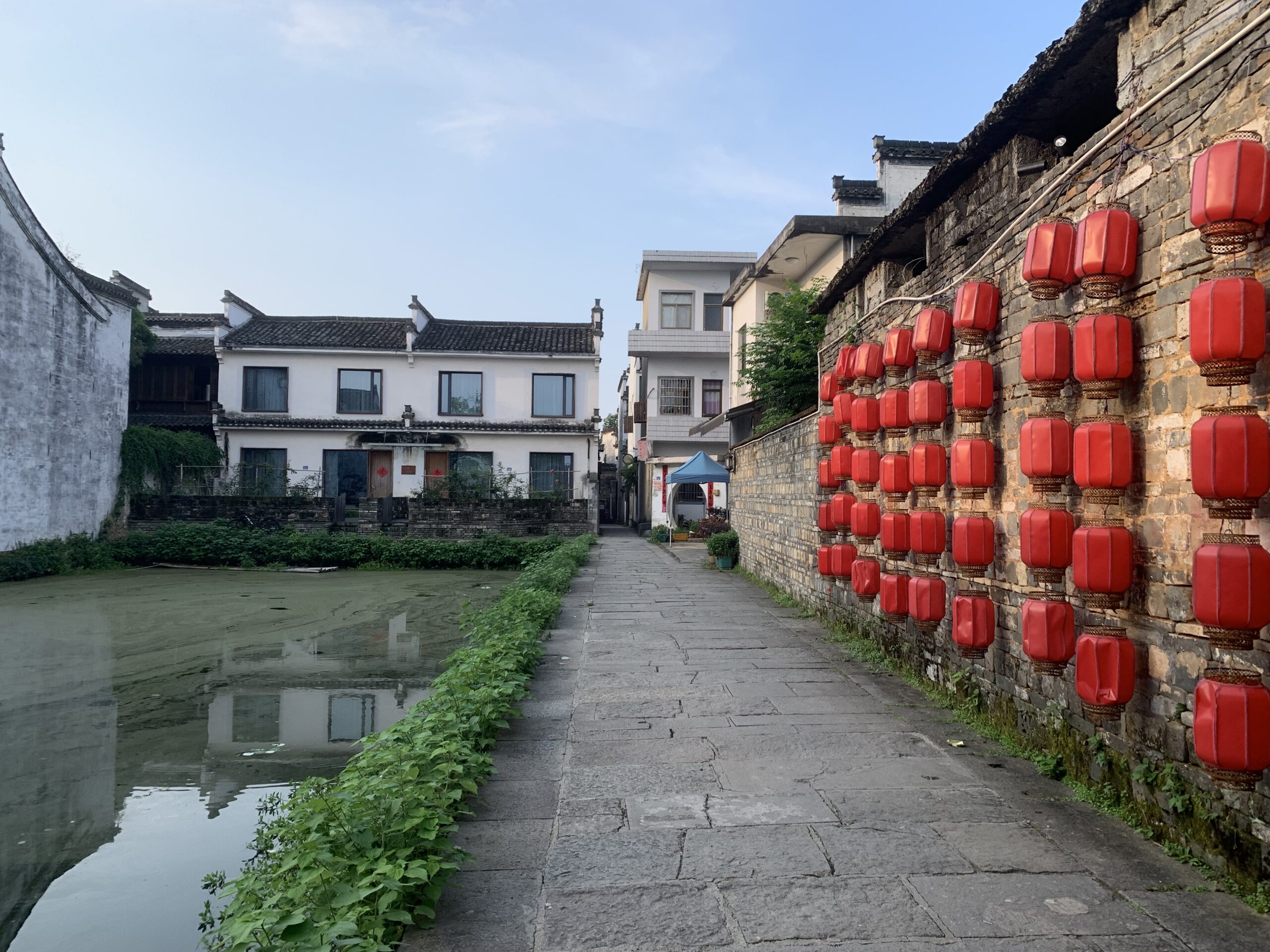 Ruelle pavée bordée d'eau et mur en pierre orné de lanternes rouges à Xixinan, à ne pas manquer pour visiter Qimen.