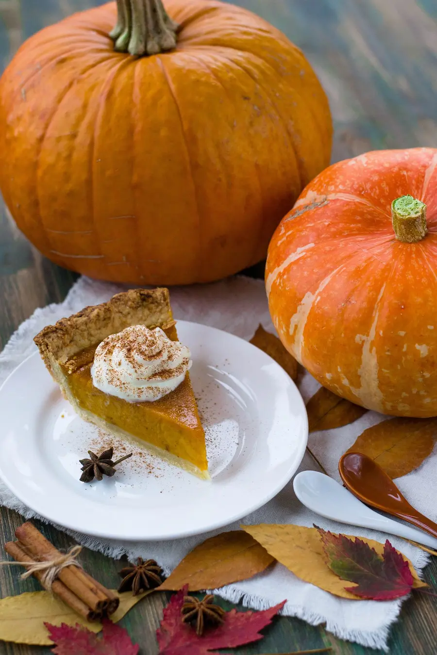 Part de pumpkin pie surmontée de chantilly et d'épices entourée de citrouilles et feuilles mortes, idéale pour un accord thé et dessert d'automne cet automne.